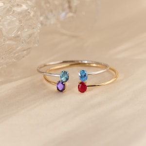 May include: Two open-ended bracelets, one silver and one gold, each adorned with two oval gemstones. The gemstones are blue, purple, and red. The bracelets are displayed on a light beige surface.