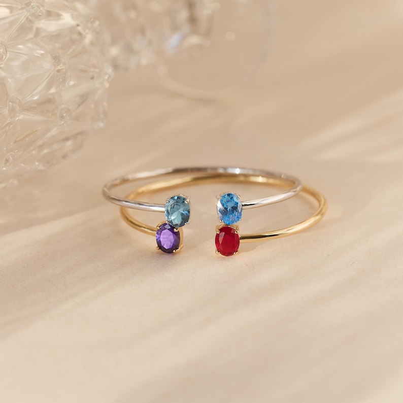 May include: Two open-ended bracelets, one silver and one gold, each adorned with two oval gemstones. The gemstones are blue, purple, and red. The bracelets are displayed on a light beige surface.