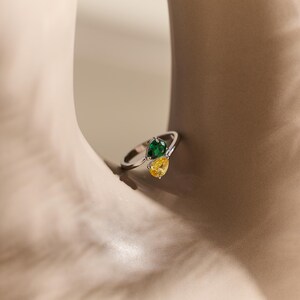 May include: A silver ring with two pear-cut gemstones, one green and one yellow. The ring has a contemporary design, with the stones set next to each other. The ring is shown against a neutral backdrop.