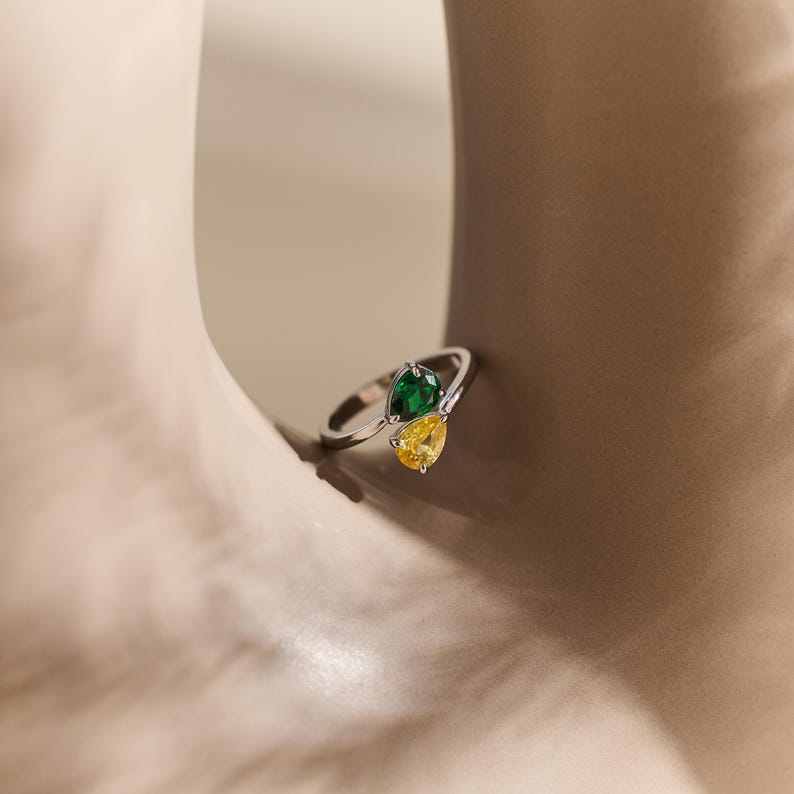 May include: A silver ring with two pear-cut gemstones, one green and one yellow. The ring has a contemporary design, with the stones set next to each other. The ring is shown against a neutral backdrop.