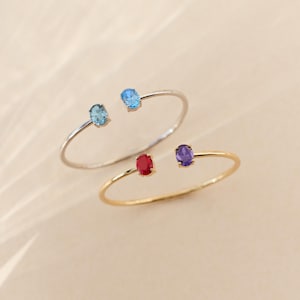 May include: Two open cuff bracelets. One is silver with blue gemstones, the other is gold with red and purple gemstones. The gemstones are oval-shaped and set at each end of the bracelet.
