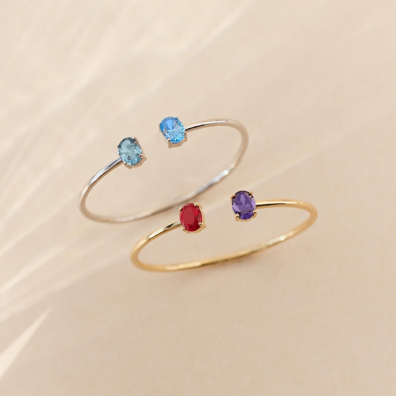 May include: Two open cuff bracelets. One is silver with blue gemstones, the other is gold with red and purple gemstones. The gemstones are oval-shaped and set at each end of the bracelet.