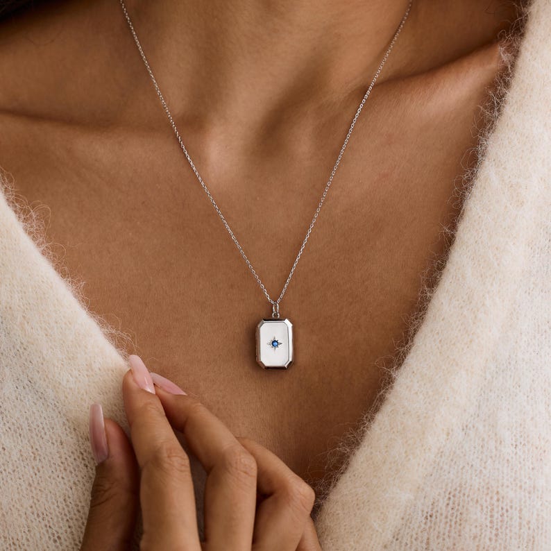 Puede incluir: Un collar de plata con un colgante rectangular. El colgante tiene un centro de esmalte blanco con un dise&ntilde;o de estrella azul. El collar lo lleva una persona.