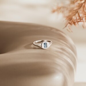 May include: A silver ring with a rectangular, light blue gemstone set in a rectangular bezel. The ring is on a neutral-toned surface, with soft lighting and a blurred background.