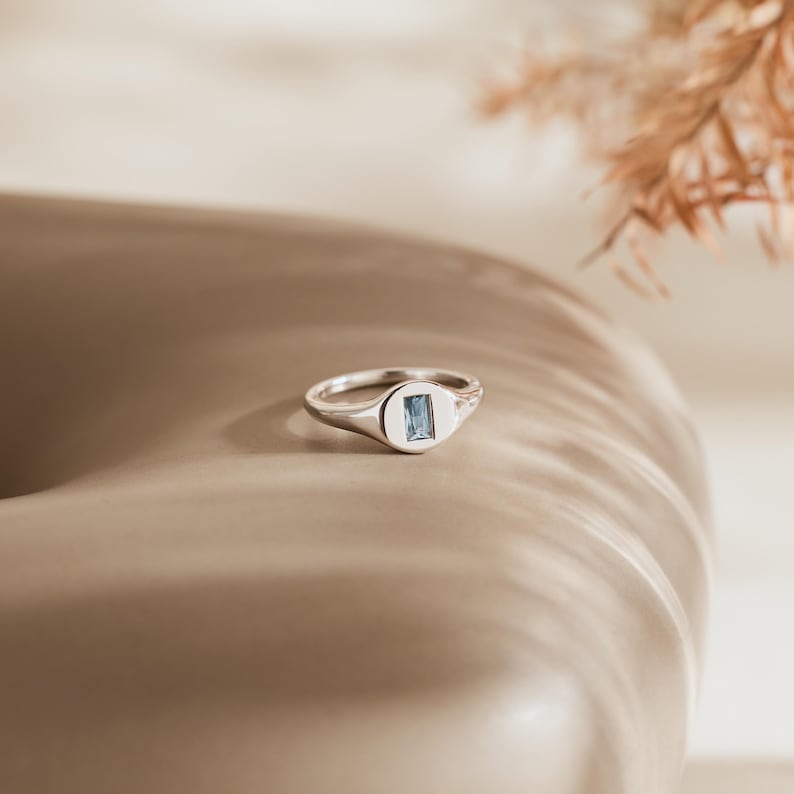 May include: A silver ring with a rectangular, light blue gemstone set in a rectangular bezel. The ring is on a neutral-toned surface, with soft lighting and a blurred background.