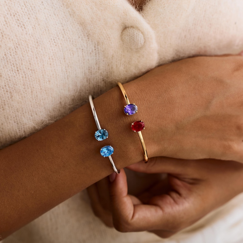 May include: Two open cuff bracelets, one silver and one gold, each adorned with oval gemstones. The silver bracelet features two blue gemstones, while the gold bracelet showcases a purple and a red gemstone. The bracelets are worn on a wrist.