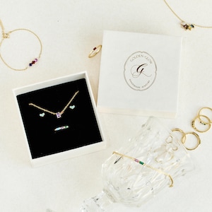 Puede incluir: Una variedad de joyas doradas, que incluyen collares, anillos y una pulsera, se exhiben sobre una superficie blanca. Tambi&eacute;n hay una caja blanca con el texto "GOLDEN GLOW HANDMADE JEWELRY". Las joyas presentan piedras preciosas de colores y detalles en forma de coraz&oacute;n.