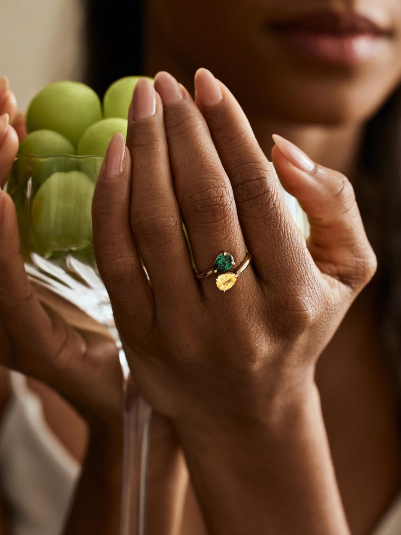 May include: A gold ring with a pear-shaped emerald and a yellow gemstone. The ring is on a hand holding a glass of green grapes. The ring's gemstones are set in a gold band.