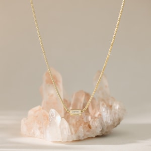 May include: A gold chain necklace with a small, engraved cylinder charm. The charm has the names "Daniel" and "James" engraved on it. The necklace is displayed on a bed of pink and white crystals.