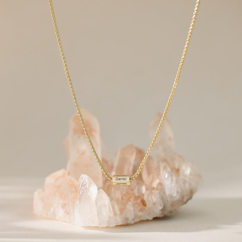 May include: A gold chain necklace with a small, engraved cylinder charm. The charm has the names "Daniel" and "James" engraved on it. The necklace is displayed on a bed of pink and white crystals.