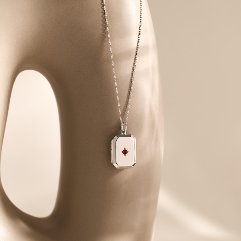 Puede incluir: Un collar plateado con un colgante rectangular con un dise&ntilde;o de estrella roja. El colgante cuelga de una cadena fina. El fondo es de tono neutro.