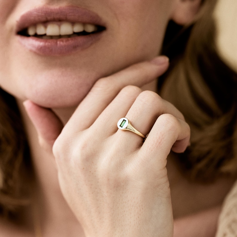 May include: A gold ring with a small, rectangular green gemstone set in a white circular setting. The ring is on a finger, with the hand resting near the face. The ring's band is thin and polished.
