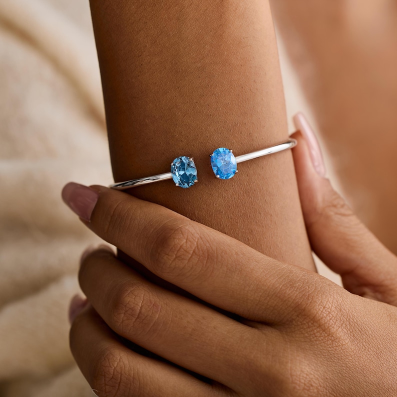 May include: A silver-toned open cuff bracelet featuring two oval-cut, light blue gemstones. The bracelet is shown on a person's wrist, highlighting the gemstones' color and the bracelet's design. The gemstones are set on either end of the bracelet.
