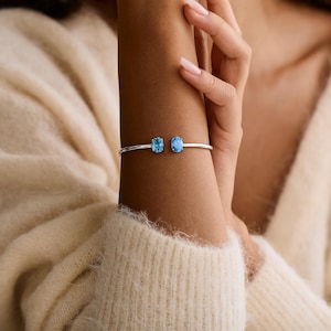 May include: A silver-toned bracelet featuring two oval-shaped, light blue gemstones. The bracelet is worn on a wrist, with the gemstones positioned side-by-side. The gemstones are approximately 0.3 inches in length.