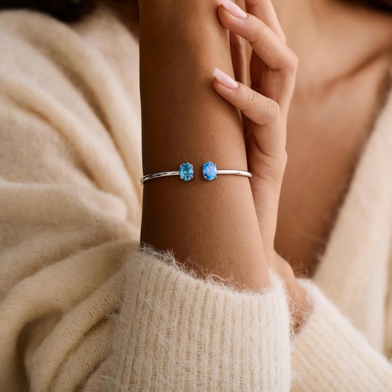 May include: A silver-toned bracelet featuring two oval-shaped, light blue gemstones. The bracelet is worn on a wrist, with the gemstones positioned side-by-side. The gemstones are approximately 0.3 inches in length.