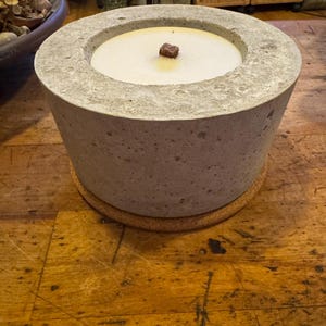 Könnte beinhalten: Ein grauer Beton-Kerzenhalter mit runder Oberseite und einem kleinen braunen Docht in der Mitte. Der Kerzenhalter steht auf einer Holzoberfläche.