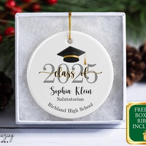 May include: A white ceramic graduation ornament with a black mortarboard, gold tassel, and the text "Class of 2025." The ornament includes the name "Sophia Klein," and "Salutatorian" and "Richland High School." It is presented in a gift box.