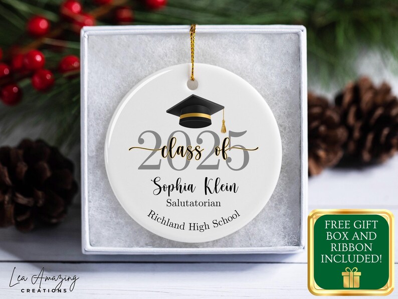 May include: A white ceramic graduation ornament with a black mortarboard, gold tassel, and the text "Class of 2025." The ornament includes the name "Sophia Klein," and "Salutatorian" and "Richland High School." It is presented in a gift box.