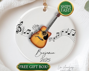 May include: White ceramic ornament with a guitar and musical notes design. The ornament features a brown and yellow acoustic guitar, black musical notes, and the name "Benjamin" with the year "2025". A gold-colored string is attached for hanging.