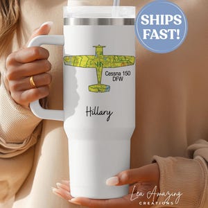 May include: A white travel tumbler with a handle, featuring a yellow and green airplane design with "Cessna 150 DFW" text. The name "Hillary" is printed below the design. A blue circle with "SHIPS FAST!" is in the background.