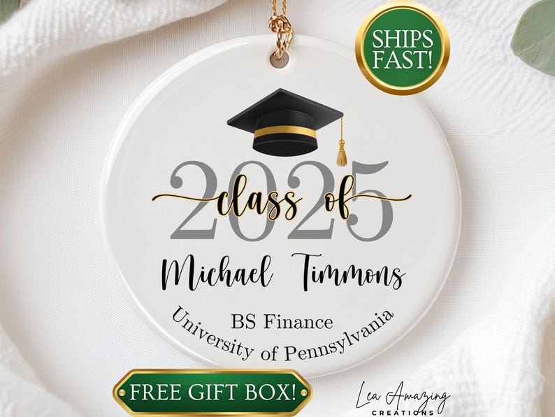 May include: A white ceramic graduation ornament with a black mortarboard and gold tassel. The ornament reads "Class of 2025" and includes a name, degree, and university. The ornament is round and has a gold hanging loop.