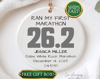 May include: White ceramic ornament with gold-colored hanging cord. The ornament features the text "RAN MY FIRST MARATHON" above the numbers "26.2" in gray. Below, it reads "JESSICA MILLER" and marathon details. A green and gold sticker says "SHIPS FAST!"