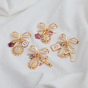 Può includere: Quattro spille a fiocco dorate con ciondoli pendenti. Ogni spilla presenta un design a fiocco e vari ciondoli, tra cui simboli religiosi, cristalli e pendenti con iniziali. Le spille sono poste su uno sfondo di tessuto bianco.