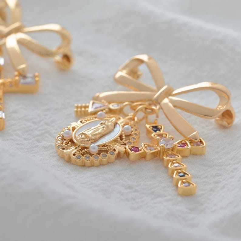 May include: Gold-toned earrings featuring a bow design. Each earring has a cross pendant adorned with colorful gemstones and a medallion with a religious figure. The earrings are set against a white background.