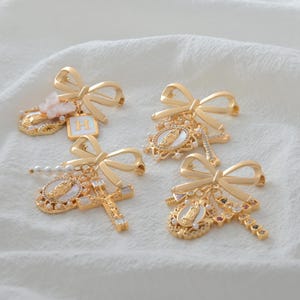 Può includere: Quattro spille a fiocco dorate con vari ciondoli. Ogni spilla presenta un design a fiocco ed è adornata con simboli religiosi, perle e pietre preziose colorate. Le spille sono esposte su una superficie bianca e strutturata.