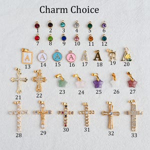 May include: A collection of gold-toned charms, including crosses, stars, and pendants with letters and colorful gemstones. The text "Charm Choice" is at the top. The charms are arranged on a white surface.