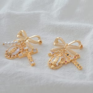 May include: Two gold-colored bow brooches with religious charms. One brooch features a cross with clear crystals, a pearl strand, and a medallion with a religious figure. The other brooch has a similar medallion and a cross with colorful gems.
