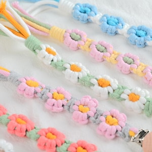 Può includere: Primo piano di diverse mollette per capelli a fiori in macramè fatte a mano in vari colori. Le mollette presentano fiori a forma di margherita in rosa, bianco, giallo e blu, con accenti verdi e grigi. Le mollette sono disposte su una superficie bianca.