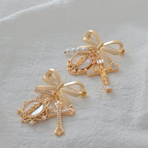 May include: Two gold-colored brooches with bow details. Each brooch features a cross, a medallion with a religious figure, and faux pearls. The brooches are set against a white, textured background.