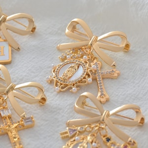 May include: Gold-toned bow brooches with religious charms. Each brooch features a bow design and a pin backing. Charms include crosses, oval pendants with religious figures, and pearl accents. The brooches are displayed on a white, textured surface.