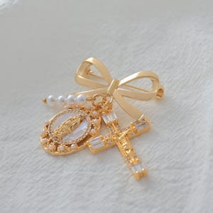 May include: Gold-toned religious brooch featuring a bow, a cross with clear crystals, and an oval pendant with a religious figure. The brooch also includes a string of pearls, all set against a white background.