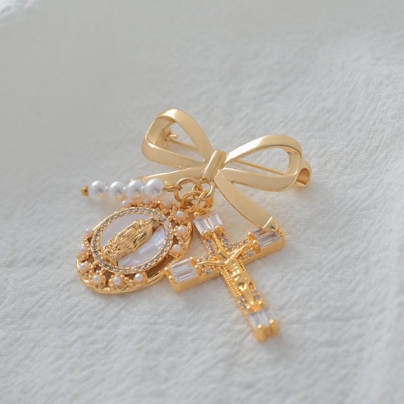 May include: Gold-toned religious brooch featuring a bow, a cross with clear crystals, and an oval pendant with a religious figure. The brooch also includes a string of pearls, all set against a white background.