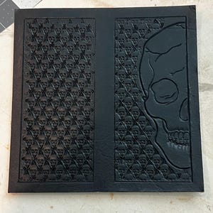 Op de afbeelding: Zwarte leren journal cover met een schedel ontwerp. De cover heeft een geometrisch patroon van kleine schedels en een grote schedel aan de rechterkant. Het journal is rechthoekig en heeft een getextureerd oppervlak.