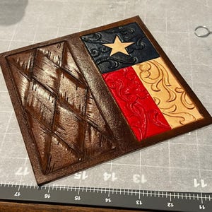 May include: Hand-tooled leather wallet featuring the Texas flag design. The wallet has a brown diamond-patterned section and a flag design with a blue top, red middle, and tan bottom. A gold star is in the blue section. The edges are brown.