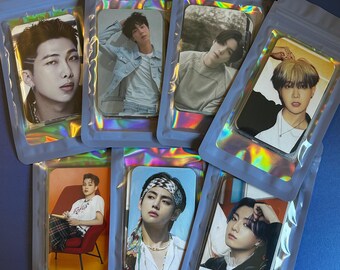 May include: A collection of seven photo cards featuring different people, each in a clear, iridescent pouch. The cards display portraits of the individuals, with varying poses and expressions. The pouches are sealed and arranged on a blue surface.