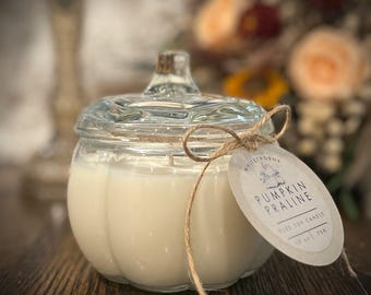 May include: A clear glass pumpkin-shaped candle jar filled with a creamy white candle. The jar has a glass lid and is tied with twine and a tag that reads "Pumpkin Praline". The candle is 10 oz / 283g.