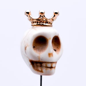 May include: A small, off-white skull figurine with a gold-colored crown. The skull has dark eye sockets and a detailed jawline. The figurine is mounted on a thin, black rod, set against a white background.