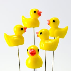 May include: Five yellow duck pins with red beaks and black eyes are arranged in a circular pattern. The pins are on thin silver stems, and the ducks are facing different directions. The background is white.