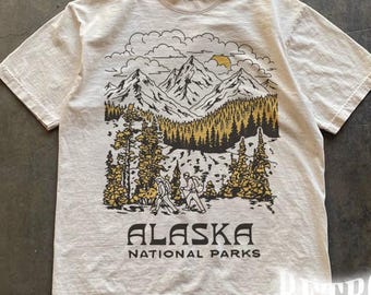 May include: Ivory t-shirt with a mountain landscape graphic and the text "ALASKA NATIONAL PARKS." The design includes mountains, trees, and hikers. The shirt has short sleeves and a crew neck. The colour is ivory.