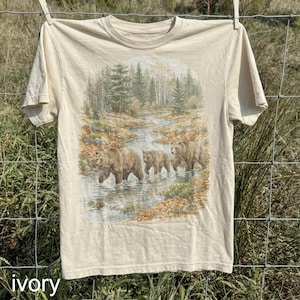 Può includere: T-shirt color avorio con un'illustrazione dettagliata di tre orsi bruni che camminano in un ruscello in una foresta. Il design include alberi, acqua e fogliame, creando una grafica a tema natura. La parola "ivory" è stampata in basso.