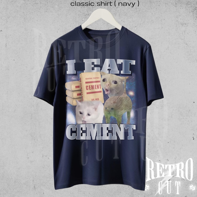 Op de afbeelding: Marineblauw t-shirt met een humoristische afbeelding van drie katten en de tekst "I EAT CEMENT". De katten worden afgebeeld met een zak cement en de tekst "CEMENT" eronder. Het shirt hangt aan een witte hanger.