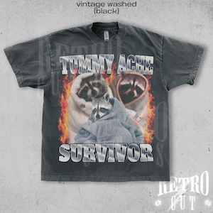 May include: A black vintage washed t-shirt featuring a graphic print of three raccoons wearing hooded sweatshirts. The text "Tummy Ache Survivor" is printed in silver on the front of the shirt.