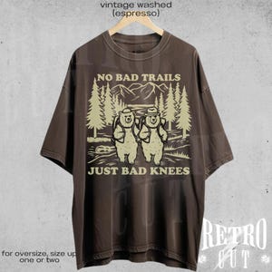 Op de afbeelding: Een bruin t-shirt met een vintage gewassen look. Het shirt heeft een afbeelding van twee beren die in een bos wandelen met de tekst "NO BAD TRAILS JUST BAD KNEES".