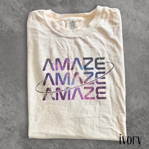 Könnte beinhalten: Elfenbeinfarbenes T-Shirt mit dem Wort "AMAZE" in Blau-, Lila- und Schwarztönen dreimal gedruckt, mit einer geschwungenen Linie. Das T-Shirt ist sauber gefaltet.