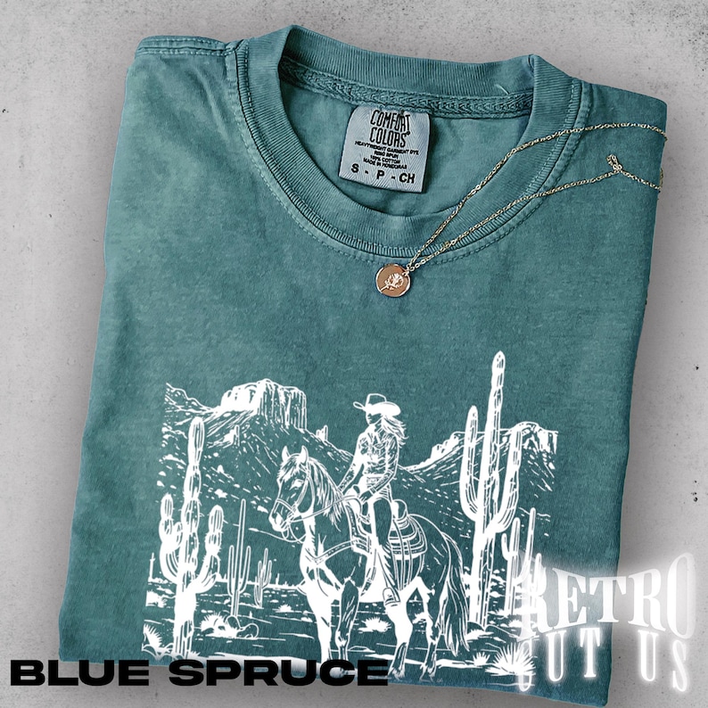 May include: Blue spruce t-shirt with a white graphic of a cowgirl on a horse in a desert landscape. The shirt has a crew neck and short sleeves. A gold necklace with a pendant is draped across the shirt. The text "BLUE SPRUCE" is at the bottom.