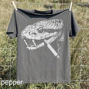 May include: A gray t-shirt with a white graphic of a snake's head smoking a cigarette. The shirt is hanging on a clothesline. The word "pepper" is printed in white at the bottom of the shirt.